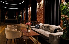 Двухуровневая event-площадка 550 м². Лофт-пространство с кирпичными стенами и деревянным полом. Мягкий серый диван, два бежевых кресла и деревянный стол с цветами и свечой. Черные занавески и круглые светильники над зоной отдыха.