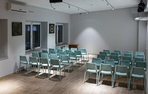 Пространство для ваших праздников. Конференц-зал с деревянным полом, расставлены ряды мятных стульев. Светлые стены, на одной из них картины. Потолочные светильники обеспечивают освещение. Есть кондиционер и окна с откосами. У стены небольшой деревянный стол.