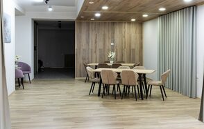 Пространство для ваших праздников. Просторное помещение с деревянным полом и панелями на стенах и потолке. Две группы столов и стульев, цветы на столах. Освещение представлено встроенными светильниками. Справа - шторы, слева - картина и пара кресел.