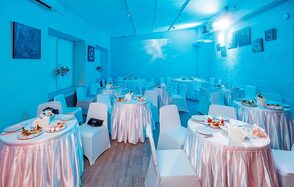 Пространство для ваших праздников. Банкетный зал с круглыми столами, покрытыми белыми скатертями, и белыми стульями. Голубое освещение. На стенах картины. Декор из цветов. Блюда с закусками на столах. Пол деревянный. Потолочное освещение.