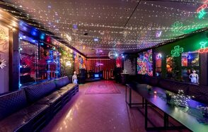 Яркий лофт для праздников. Караоке-зал с диванами вдоль стен, деревянные столы, зеркальные панели, яркие неоновые и светодиодные украшения на стенах, цветной ковёр по центру, отражения огней на потолке, тёмная цветовая гамма интерьера.