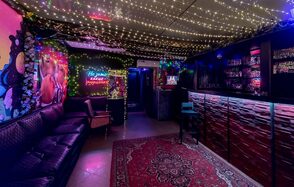 Яркий лофт для праздников. Клубное пространство с черным кожаным диваном, барной стойкой, полками с напитками. Потолок украшен гирляндами, стены с неоновыми знаками и граффити. На полу ковёр с орнаментом. Освещение приглушенное, неоновое и гирляндное.