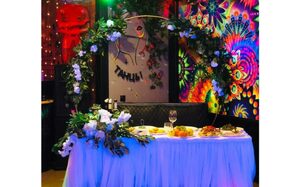 Яркий лофт для праздников. Светлое помещение с декорированным столом, покрытым белой скатертью, украшенным цветочной аркой. На столе еда и напитки. На стене яркие рисунки и надпись "Танцы!" Окружение подсвечено разноцветными огнями.