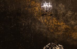 Лофт со своим двориком. Потолочный светильник в виде клетки подвешен на фоне текстурированной темной стены с золотистыми акцентами. Справа внизу металлический декоративный элемент с абстрактным узором. Освещение теплое.