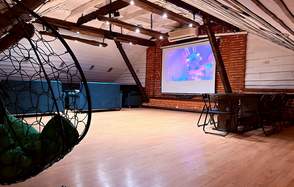 Лофт-мансарда . Атмосферное лофт-пространство с деревянным полом и скошенным потолком, кирпичная стена. Есть гамаки, кожаные диваны, черный стол с металлическими стульями. Установлен проектор. Подвесное освещение, деревянные балки, светильники на потолке.