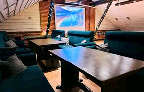 Лофт-мансарда . Лофт-пространство с кирпичной стеной и наклонным потолком. Расстановка из деревянных столов и темно-синих диванов с подушками. На стене проектор с изображением. Потолочные балки украшены гирляндами. Освещение мягкое, теплое. Пол деревянный.