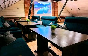 Лофт-мансарда . Лофт с кирпичными стенами и деревянными панелями. Цифровой экран на стене, темные деревянные столы, темно-зеленые кресла с подушками. На столах горшки с зелеными растениями. Зафиксировано декоративное освещение. Светлый деревянный пол.