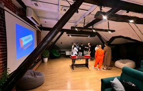 Krisha loft (Крыша Лофт). Лофтовой пространство с деревянным полом и кирпичными стенами. На стене висит большой экран. Мебель: диваны, кресло-мешок, стол с плоской столешницей. Несколько человек стоят у стола. Освещение подвесными лампами, интерьер выдержан в спокойных тонах.