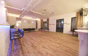 Loft Lodge(Лофт Лодж) 9. Просторное помещение с большим количеством открытого пространства, деревянный пол, белые кирпичные стены, высокие стулья у стойки, стулья у окна, круглая люстра, светлая мебель, разноцветная подсветка, большие окна с занавесками, настенные часы, декоративные огни.