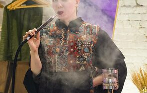 Лофт из белого кирпича в центре. Человек курит кальян в помещении. На стене сзади белая кирпичная кладка. Справа стоит кальян с металлическими элементами. На заднем плане видна стойка с одеждой. Помещение освещено мягким светом.