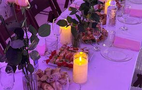 Лофт из белого кирпича в центре. Белый стол с розовой скатертью, сервирован стеклянной посудой и столовыми приборами. На столе розы в вазах, декоративные свечи, разложены закуски. Складные деревянные стулья вокруг. Освещение тёплое, фиолетовое.
