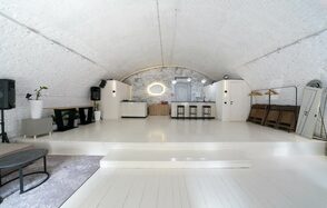 Brodniki Loft (Бродники Лофт). Лофт из белого кирпича. Арочное помещение белого цвета с деревянным полом и ступенькой. На заднем плане барная стойка, стулья, колонки и акустическое оборудование. Декоративное зеркало на кирпичной стене. Слева стулья и журнальный столик. Освещение встроенное.