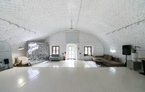 Brodniki Loft (Бродники Лофт). Лофт из белого кирпича. Белый сводчатый зал с деревянным полом, мягкие серые и коричневые диваны, напольная лампа, стена с надписью, три окна, дверной проем, пожарный щит. Потолочное освещение и подвесная небольшая лампа. Стены и потолок из кирпича, белый цвет.