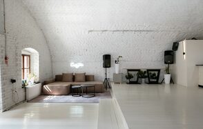 Brodniki Loft (Бродники Лофт). Лофт из белого кирпича. Арочное пространство с белыми кирпичными стенами и светлым деревянным полом. Коричневый диван с подушками на ковре, рядом с круглым столиком. По обеим сторонам украшения в виде растений в белых горшках. Два черных динамика. Окно с белыми рамами слева.
