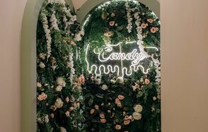 Лофт Кэнди. Арка с декором из зелени и цветов, неоновая вывеска с текстом "Candy" на зелёной стене с растениями. Цветы белые и розовые, стенка покрыта плотной листвой. Освещение от вывески; рамка арки светлого оттенка.