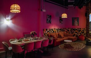 Лофт Гламур. Розовая комната с обеденным столом и стульями, сервировка на столе, оранжевый угловой диван, круглый столик, декор с леопардовым принтом, настенные картинки, подвесные светильники с абажурами, светлое точечное освещение, пол темного цвета.