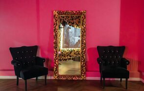 Лофт Гламур. Пространство с ярко-розовыми стенами, с двумя черными креслами с пуговицами по бокам от зеркала в золотистой раме с узором. Зеркало отражает зал с принтом под зебру, освещение теплое, пол из темного дерева.