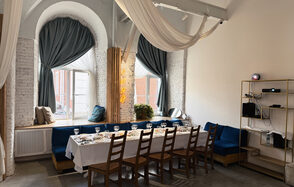 Малый светлый лофт для мероприятий. Просторная комната с белыми кирпичными стенами, высокими сводчатыми окнами с синими шторами. Длинный стол с белой скатертью, оформленный стеклянной посудой, окружён деревянными стульями. Слева синее мягкое сиденье, справа стеллаж с техникой. Светлый потолок с тканевым декором.