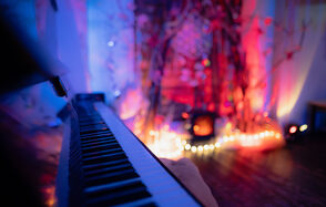 Лес. Театральный лофт с верандой, сценой и вторым ярусом. Пианино крупным планом с боковой стороны, на заднем плане декор из веток с цветными огнями и гирляндами. Мягкое разноцветное освещение в синих и красных тонах. Пол тёмного оттенка, стены светлые.