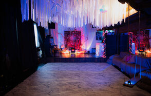 Лес. Театральный лофт с верандой, сценой и вторым ярусом. Просторное помещение с белым узорчатым полом и темными стенами, украшено длинными белыми лентами под потолком. По периметру разнообразная мебель, включая диван и декорации с разноцветной подсветкой. На сцене красочная инсталляция. Свет приглушен.