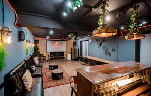 Пространство для любых мероприятий с верандой и грилем. Просторное помещение с мягкими диванами вдоль стены, барной стойкой и длинным деревянным столом. Настенное освещение в виде ламп, подвесные светильники с растениями, экран с проектором. Декор из зелени и светлое деревянное покрытие пола. Стены синие.