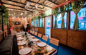 Пространство для любых мероприятий с верандой и грилем. Длинный обеденный стол с белыми тарелками и стеклянной посудой, деревянные стулья, свисающие растения, зелёные шторы на окнах, деревянные панели, подвесные светильники, имитация травы на потолке, дартс на стене, уютный интерьер.