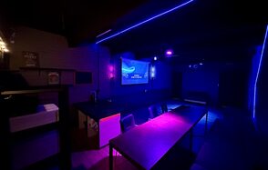Kahovka loft (Каховка Лофт). Неоновый лофт-зал. Тёмная комната с неоновым фиолетовым освещением, длинный стол с креслами, телевизор на стене, мягкий уголок вдоль стены, полки с декором, детали из кирпича, надпись на экране.