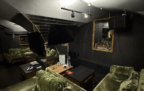 Shantaram(Шантарам). Комната с двумя мягкими зелеными диванами, кофейным столиком, черными стенами с золотыми рамами и зеркалами. Потолок с проектором и подвесными светильниками. На столике элементы декора и устройства. Темное освещение.