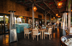 Soho Country Club (Сохо Кантри Клаб). Открытая терраса с деревянными полами и потолком, украшена висящими светильниками. Расставлены столы и стулья из светлого дерева. На заднем плане стеклянные двери, по бокам уличные обогреватели. Декор в теплых тонах с зелеными элементами.