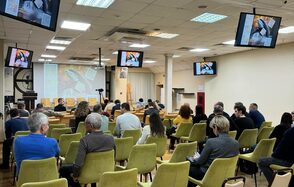 Зал 9. Просторный конференц-зал до 200 человек в Академии Кантаты. Конференц-зал с рядами зеленых стульев, оборудован проектором и несколькими экранами, показывающими изображения. Потолок с встроенными светильниками. Люди сидят, некоторые с блокнотами. Белые стены, деревянный пол, хорошее состояние.