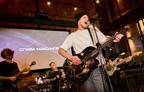 Лофт для мероприятий с гольф-симулятором. Зал с кирпичными стенами, сценой и музыкальными инструментами, включая гитары и барабаны. Мужчина поет в микрофон, держа гитару. На заднем плане экран с текстом. Освещение точечное, теплый свет.