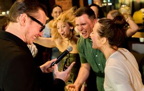 Лофт для мероприятий с гольф-симулятором. Группа людей веселится в помещении, один человек держит микрофон. Видна кирпичная стена, задний план размытый. Яркое освещение, тёплые цвета, присутствует барная стойка.