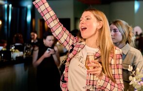 Лофт для мероприятий с гольф-симулятором. Группа людей, одетых в современную одежду, находятся в помещении с барной стойкой. Освещение тёплое, присутствуют элементы декора. Один человек держит бокал. Цветовая гамма включает тёмные и светло-зелёные оттенки.