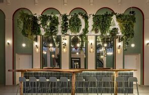 Viola Loft (Виола Лофт). Просторный и светлый лофт с панорамным видом в парк. Барное пространство с длинной деревянной стойкой и серыми стульями. Вверх украшен зеленью и светильниками. На заднем плане большие зеркала и арочные ниши. Стены светло-зеленые, акцент выполнен красной аркой. Освещение теплое.