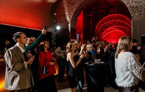 Polygon(Полигон). Зал Кухня. Арочное кирпичное пространство с приглушённым освещением в красных тонах. Группы людей стоят, держа бокалы. Высокие круглые столы, декоративные световые панели на стенах. Атмосфера торжества или события.