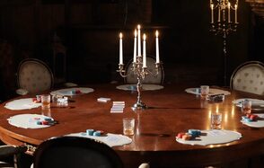 Polygon(Полигон). Зал Апартаменты. Большой круглый деревянный стол с нотками ар-деко, окруженный классическими стульями с узором. На столе расположены канделябры с зажженными свечами, карточки, стаканы и игровые фишки. Освещение теплое, приглушенное. Цвета темные и нейтральные.