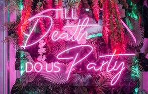 Rubin Loft (Рубин Лофт). Оушен Драйв. Неоновая надпись "Till Death Do Us Party" розовым светом на фоне зелени и искусственных цветов, включая розовые и красные элементы. Освещение создает яркий контраст между светом и тенями, добавляя атмосферность.