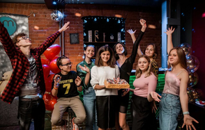 Клубный лофт на набережной в центре Москвы. Кирпичная стена, барная стойка с подсветкой, светильники, диско-шар, серые и зелёные цвета, шарики, праздничные украшения, группа людей позирует, держат торт.