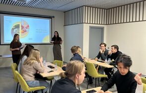 Зал 6. Просторный и светлый конференц-зал до 50 человек в Академии Кантаты. Конференц-зал с тремя длинными деревянными столами и зелеными стульями, светло-серые стены с декоративными полосами, потолок с деревянными панелями. Проектор с экраном, на экране текст и изображение. Несколько людей сидят за столами, двое стоят у экрана.