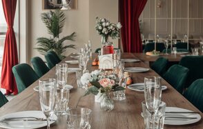 Зал Олд Мани. Лофт в индустриальном стиле. Большой деревянный стол, сервированный стеклянной посудой и белыми тарелками. По центру цветочные композиции. Мягкие зеленые стулья, красные шторы, зеленые растения. Светлая стена с картинами. Панели на заднем плане.