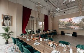 Зал Олд Мани. Лофт в индустриальном стиле. Просторное помещение с высоким потолком, длинный обеденный стол с зелёными стульями, сервированная посуда, декоративные элементы, светлый деревянный пол, красные шторы, большие зеркала, проекционный экран, зелёные растения, подвесные люстры.