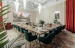 Зал Олд Мани. Лофт в индустриальном стиле. Просторное помещение с длинным деревянным столом, окружённым зелёными стульями. Подвесные люстры, красные шторы, ковёр с узором. На стене большая картина и светильник. Растения в горшках, сервировка стола с тарелками и бокалами. Светлые стены и потолок.