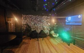 Status Loft(Статус Лофт). Лофт со сценой и караоке. Типичный Питер. Тёмное помещение с деревянным полом, на стене телевизор с видеоконтентом. Справа зелёный световой эффект. Слева диван и круглый стол. На задней стене фольгированная декорация. Несколько ламп, разноцветное освещение.