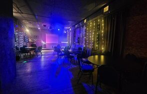 Status Loft(Статус Лофт). Лофт со сценой и караоке. Типичный Питер. Темное помещение с круглым столами и стульями. На заднем плане сцена с экраном и диджейским пультом. Праздничное освещение: гирлянды на стенах, разноцветная подсветка. Темные стены и пол. Настенная кирпичная кладка.