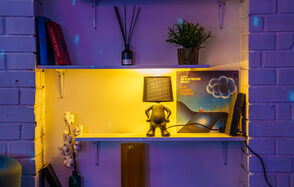 Лофт с террасой на территории Измайловского Вернисажа. Ниша в белой кирпичной стене с двумя белыми полками. На верхней: книги, ароматные палочки, растение. На нижней: лампа с фигурной ножкой, пластинка, книги. Освещение желтоватое, мебель белая. Есть растение и декоративные элементы.