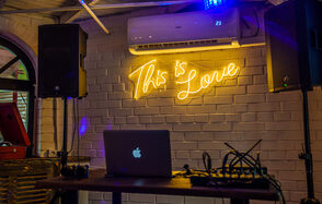 Лофт с террасой на территории Измайловского Вернисажа. Лофт-пространство с кирпичными стенами, белого цвета. На стене неоновая надпись "This is Love". Установлены аудиоколонки и кондиционер. Небольшой деревянный стол с ноутбуком и звуковым оборудованием. Мягкое освещение.