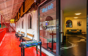 Лофт с террасой на территории Измайловского Вернисажа. Коридор с красным полом, скамейками и настольным футболом. Вывеска "Vinyl loft" на стекле. За дверью помещение с черным полом, диваном, креслом и подушками. Окна с арочными рамами, яркое освещение.