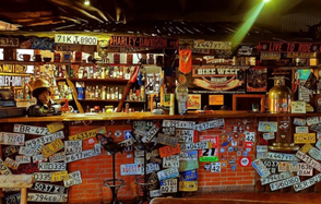 Fort Loft(Крепость лофт). Лофт в байкерском стиле. Бар с кирпичной стойкой, украшенной автомобильными номерами и подставками для ног. За стойкой полки с бутылками и декор из вывесок и флагов. На стойке статуэтка в виде шлема. Потолок деревянный, освещение тёплое, приглушённое.