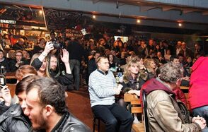 Fort Loft(Крепость лофт). Лофт в байкерском стиле. Бар с деревянными столами и стульями, значительная группа людей, сцена или подиум отсутствуют. В задней части виден бар с полками и вывеской "Sturgis." Тусклое освещение, металлический потолок, стены украшены постерами.