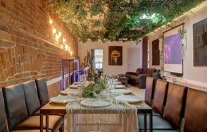 Лофт Семеновский. Лофт-пространство с кирпичной стеной. Длинный стол с сервировкой для гостей, кожаные стулья. Подвесной декор из зелени и гирлянды. На стене проектор с изображением. В зоне отдыха диваны и декор. Потолок металлический.